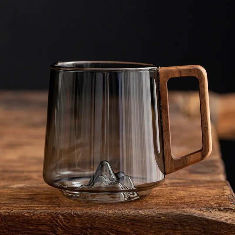 Artisan borosilicate glass mug with unique mountain base and ergonomic wooden handle for coffee or tea.