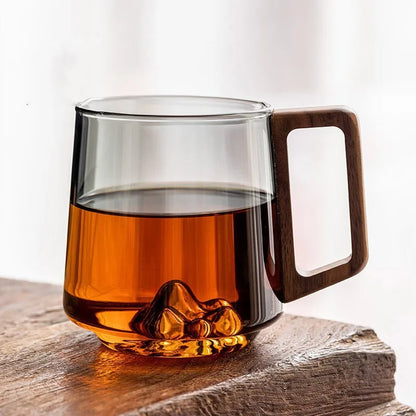 Clear borosilicate glass coffee mug featuring a sculptural mountain base & natural wood handle, perfect for home decor.